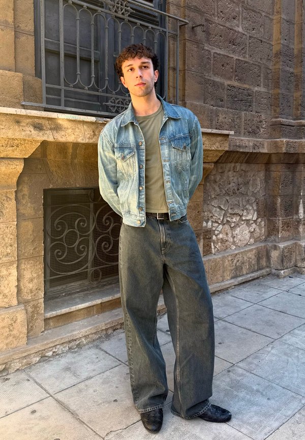 Bărbat tânăr purtând o jachetă din denim și blugi cu picioare largi, stând pe un paviment din piatră lângă un zid de piatră texturat cu grilaj metalic la fereastră.
