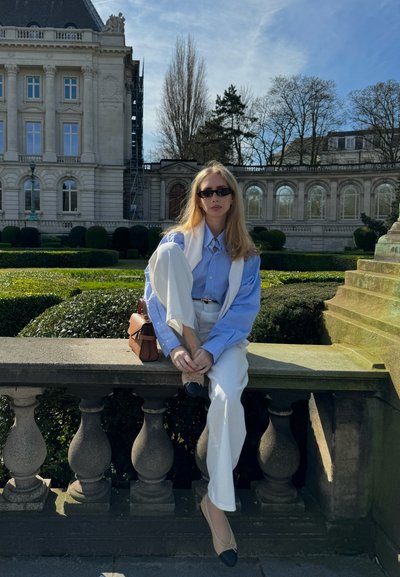 Camisa de botones azul claro, pantalones blancos, bailarinas negras y un bolso marrón. Posando sobre una barandilla de piedra con un fondo de vegetación y un edificio.