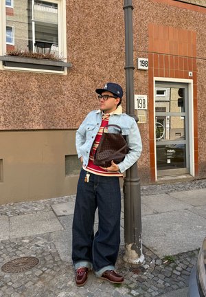 Hombre con chaqueta vaquera, camisa de rayas, vaqueros anchos y gorra, sosteniendo una bolsa marrón, apoyado en un farol en una calle urbana.