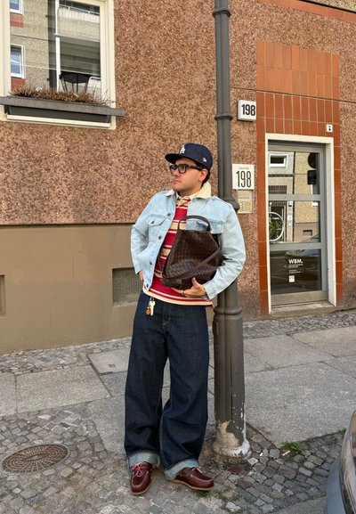 Hombre con chaqueta vaquera, camisa de rayas, vaqueros anchos y gorra, sosteniendo una bolsa marrón, apoyado en un farol en una calle urbana.