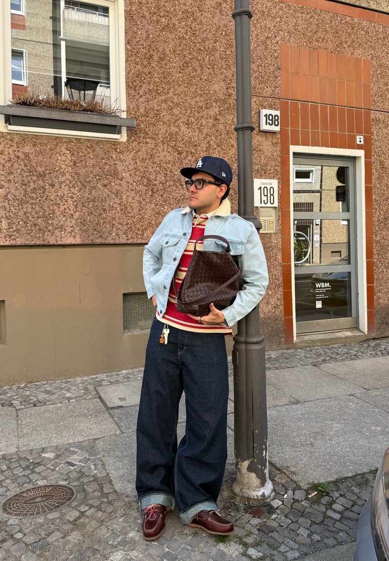Homme portant une veste en jean, une chemise rayée, un jean large et une casquette, tenant un sac marron, appuyé contre un lampadaire dans une rue urbaine.