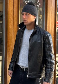 Férfi fekete sapkával, fekete bőrkabátot visel, felpattintott gallérral, szürke ing és fekete farmer áll egy faajtó mellett.