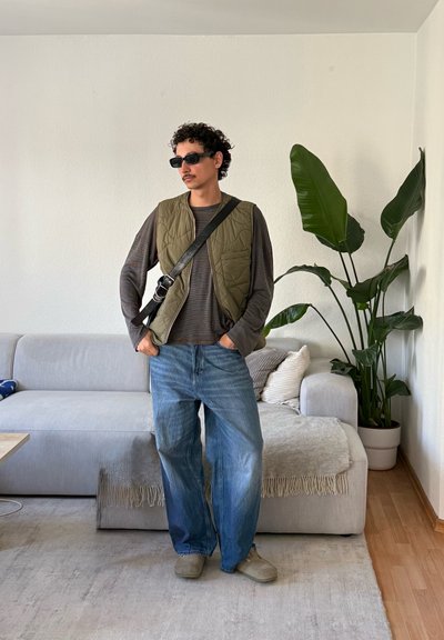 Hombre con gafas de sol, chaleco acolchado verde, camisa de manga larga, vaqueros holgados y bolso cruzado de pie en una sala de estar con sofá gris y planta grande.