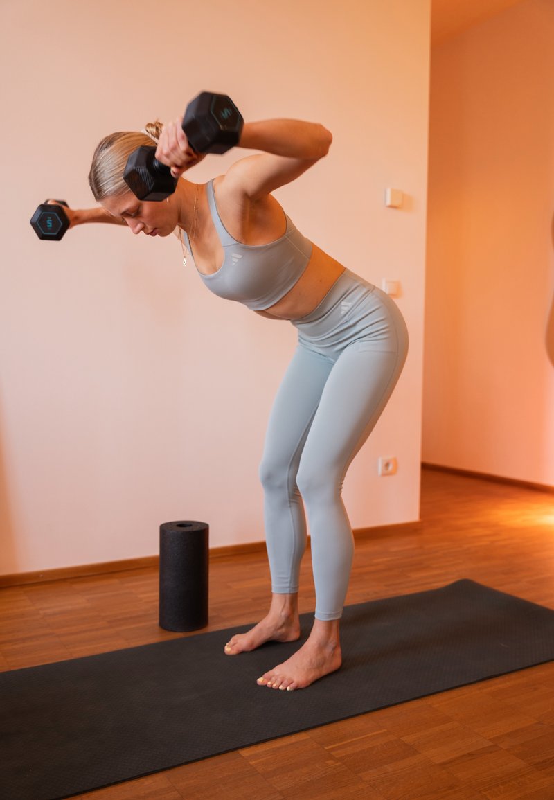 Femme en tenue de sport gris clair effectuant un reverse fly avec haltères en position penchée sur un tapis noir à l'intérieur, avec un rouleau en mousse à proximité.