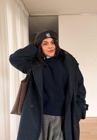 Jeune femme portant un béret noir, un pull marine, un manteau noir, une jupe plissée grise et un sac à bandoulière marron, à l'intérieur, à côté de rideaux blancs.