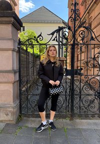 Musta lyhyt takki, mustat leggingsit ja mustat Nike-tennarit; pitäen pientä mustaa laukkua, jossa on valkoisia pisteitä, koristeellisen portin edessä.