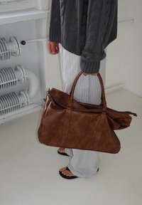 Mala de viagem em couro castanho com duas alças, parte superior com fecho éclair e um interior espaçoso. Apresenta uma superfície texturizada e um design angular.