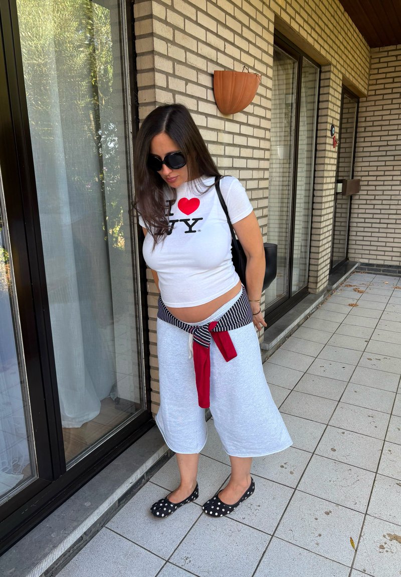 Femme portant des lunettes de soleil, un t-shirt blanc "I love NY", un short gris clair, et des ballerines noires à pois, debout sur un balcon carrelé à côté de portes vitrées.