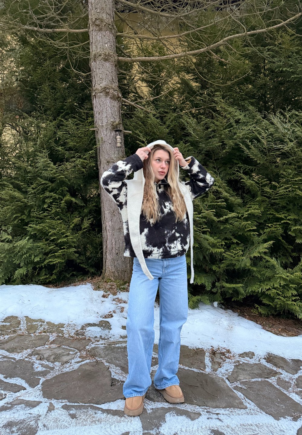 Jeune femme ajustant une capuche de couleur claire en plein air sur un chemin de pierre enneigé, vêtue d'un pull à motifs noir et blanc et d'un jean bleu clair.