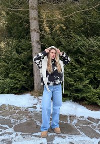 Jeune femme ajustant une capuche de couleur claire en plein air sur un chemin de pierre enneigé, vêtue d'un pull à motifs noir et blanc et d'un jean bleu clair.