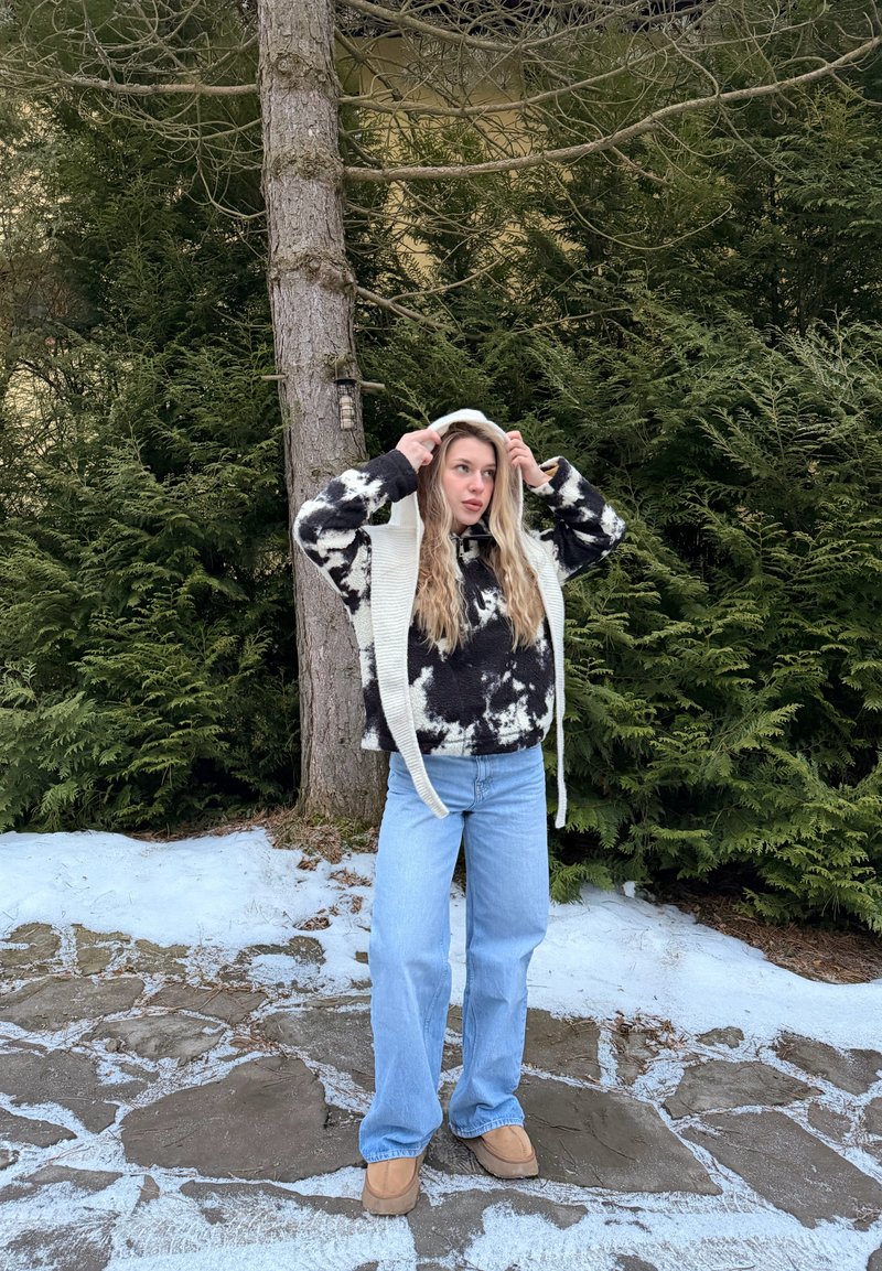 Νεαρή γυναίκα που προσαρμόζει το ανοιχτόχρωμο καπέλο της σε εξωτερικό χώρο σε χιονισμένο μονοπάτι από πέτρα, φορώντας πουλόβερ με ασπρόμαυρο σχέδιο και ανοιχτό μπλε τζιν.