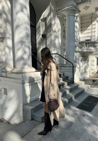 Ανοιχτό μπεζ παλτό τρέντς, μαύρο μίνι φόρεμα, μαύρες μπότες μέχρι το γόνατο με尖尖 μπροστινές μύτες και μια μικρή σκούρα τσάντα χειρός; στέκεται σε πέτρινα σκαλοπάτια με σκιές.