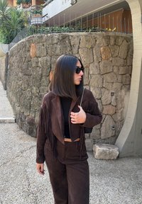 Brązowa zamszowa kurtka z frędzlami, zestawiona z pasującymi spodniami. Tekstura jest miękka, a stylizację uzupełnia czarna bluzka i okulary przeciwsłoneczne.