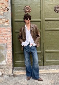 Veste en cuir marron sur une chemise blanche, associée à un jean évasé et des chaussures marron. Lunettes de soleil portées, debout contre une porte verte texturée.