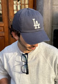 Boné de beisebol azul-marinho com o logo "LA" branco, material de algodão, aba curva, usado por uma pessoa com um sweatshirt cinza, óculos repousando na gola.