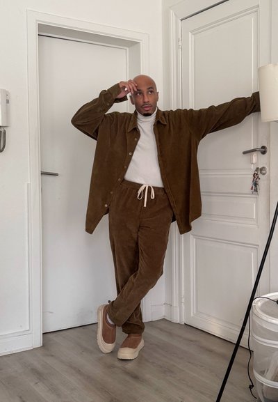 Hombre apoyado contra una puerta blanca, vistiendo chaqueta y pantalones de pana marrón, zapatos beige y un suéter de cuello alto blanco dentro de una habitación.