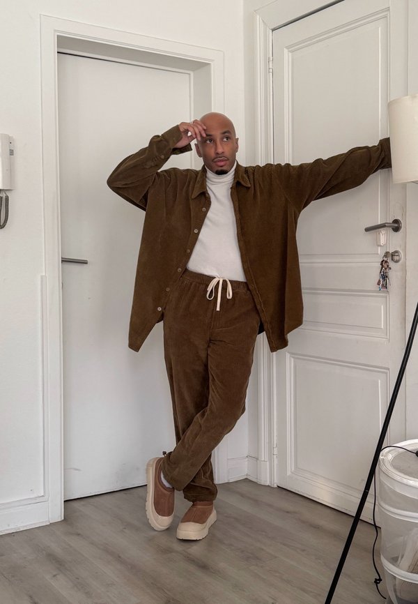 Vīrietis pieslējies pie baltām durvīm, valkājot brūnu kordurojas žaketi un bikses, bēšas kurpes un baltu augstu apkakli telpā.