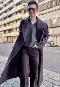 Svart lang frakk over en grå genser, stylet med mørke bukser. Store solbriller fullfører stilen. Urban bakgrunn med moderne arkitektur.