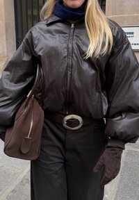 Braune gesteppte Bomberjacke mit weitem Kragen, kombiniert mit schwarzen Hosen, einem dicken marineblauen Schal, einer großen braunen Handtasche und einer runden metallischen Gürtelschnalle.