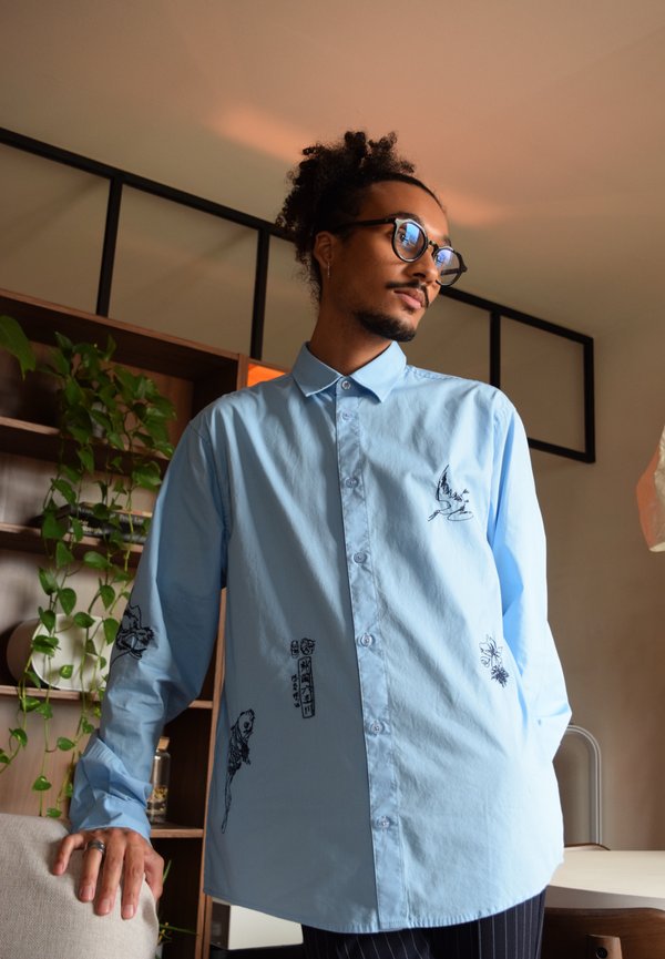 Camicia button-up azzurro chiaro con maniche lunghe, caratterizzata da motivi neri ricamati di animali e disegni sul davanti, realizzata in un tessuto liscio.