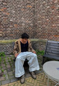 Osoba siedzi na krześle, ubrana w beżową kurtkę z futrzanym wykończeniem, czarny top na ramiączkach i szerokie białe spodnie. Widać brązowe buty; w tle ceglana ściana.