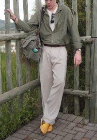 Jeune homme en pantalon beige, chemise verte et chaussures jaunes appuyé sur une clôture en bois, portant un sac en toile avec une canette de boisson, en extérieur.