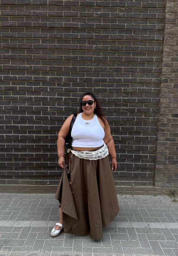 Femme portant des lunettes de soleil, un crop top blanc, une longue jupe marron avec des détails en dentelle, des chaussures blanches, debout contre un mur de briques sombre.