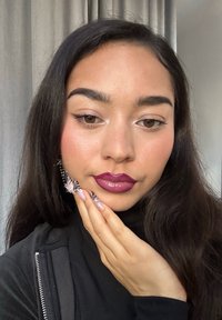 Porträt einer Person mit langen, dunklen Haaren, die einen schwarzen Rollkragenpullover trägt. Sie hat glänzend burgunderrote Lippen und kunstvoll gestaltete Fingernägel mit Mustern.