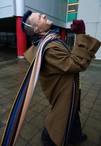 Abrigo marrón a cuadros con mangas grandes, bufanda a rayas de colores y auriculares. Suelo mojado visible en el fondo.