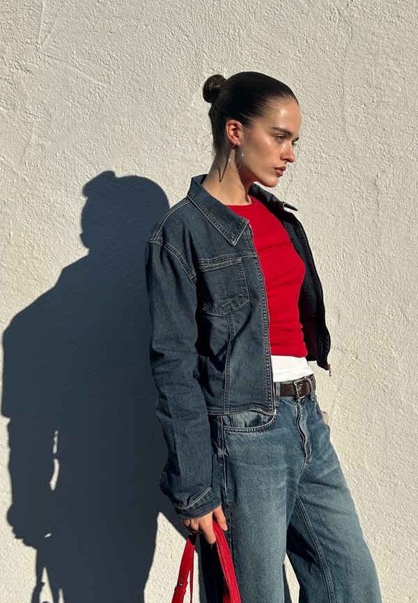 Veste en jean portée sur un haut rouge avec des accents blancs, jean bleu, et un sac à main rouge. Cheveux longs et raides ; boucles d'oreilles créoles. Ombre projetée sur un mur blanc.