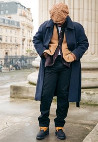 Σκούρο μπλε μάλλινο παλτό πάνω από κα�φέ γούνινη επένδυση και μωβ πουκάμισο, συνδυασμένο με μαύρο παντελόνι και καφέ μπότες με πορτοκαλί κορδόνια. Καφέ κασκέτο με καρό σχέδιο.