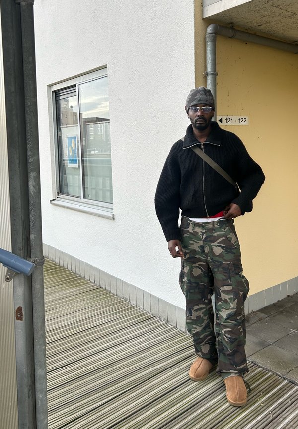 Maglione nero a maglia, pantaloni cargo mimetici con molte tasche, pantofole marroni, occhiali da sole, in piedi su un pavimento a righe vicino a una parete gialla.