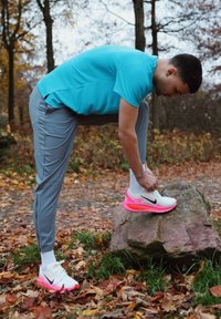 Chaussures de course Nike blanches et roses avec un détail de swoosh visible ; pantalon de sport gris et chemise bleue ; homme attachant ses chaussures sur un rocher.