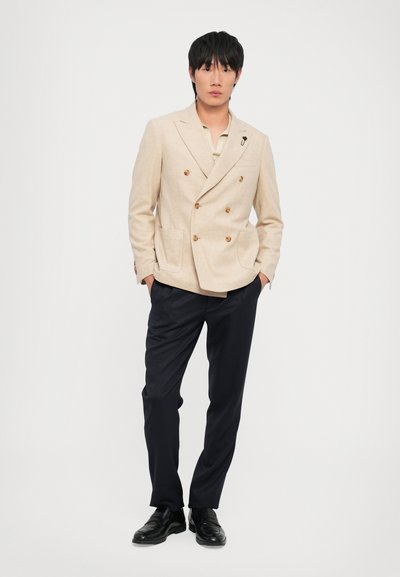 Blazer beige de doble botonadura con botones marrones, combinado con pantalones azul marino oscuros y mocasines negros. Tejido suave, ajuste entallado.
