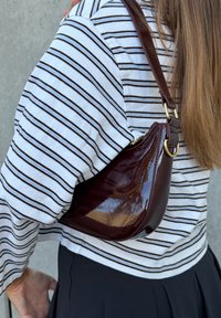 Geantă din piele lacuită burgundy cu formă curbă, accesorii aurii și o singură bretea, purtată peste un tricou cu mâneci lungi în dungi.