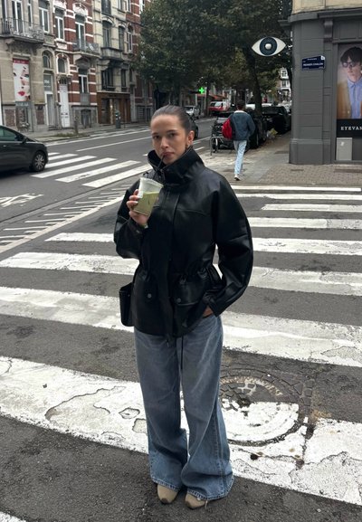 Chaqueta de cuero negra con un cuello alto, llevada sobre unos jeans azules sueltos. Sosteniendo una bebida verde, de pie en un paso de cebra a rayas.