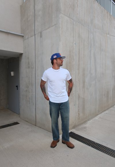 Hombre con tatuajes que lleva una camiseta blanca, vaqueros, zapatos marrones y gorra azul, apoyado contra una pared de hormigón, mirando de lado.
