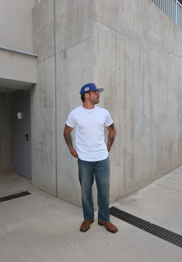 Mand med tatoveringer iført hvid t-shirt, jeans, brune sko og blå kasket, står op ad betonvæg og kigger til siden.