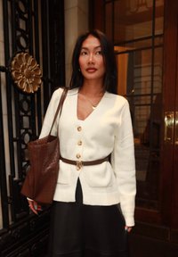 Žena v bílém kardiganu na knoflíky s hnědým opaskem a koženou taškou přes rameno stojí uvnitř u ozdobné kovové brány a skleněných dveří.