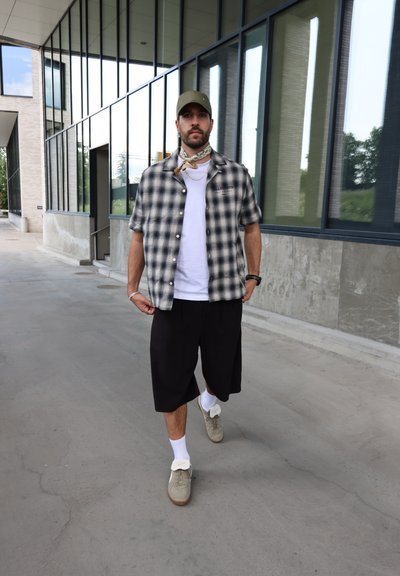 Camisa a cuadros en blanco y negro de manga corta sobre una camiseta blanca, pantalones cortos negros hasta la rodilla, zapatillas grises y una gorra verde con un pañuelo.