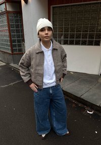 Veste bomber à carreaux marron et crème, polo blanc, jean en denim bleu surdimensionné et bonnet blanc, debout dans la rue.
