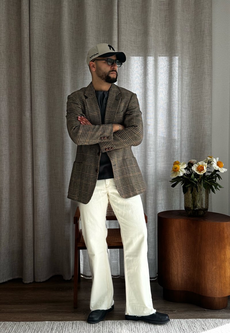Homme portant un blazer à carreaux, pantalon blanc, chaussures noires, lunettes de soleil et casquette, debout les bras croisés à côté d'une table en bois avec un vase de fleurs blanches.