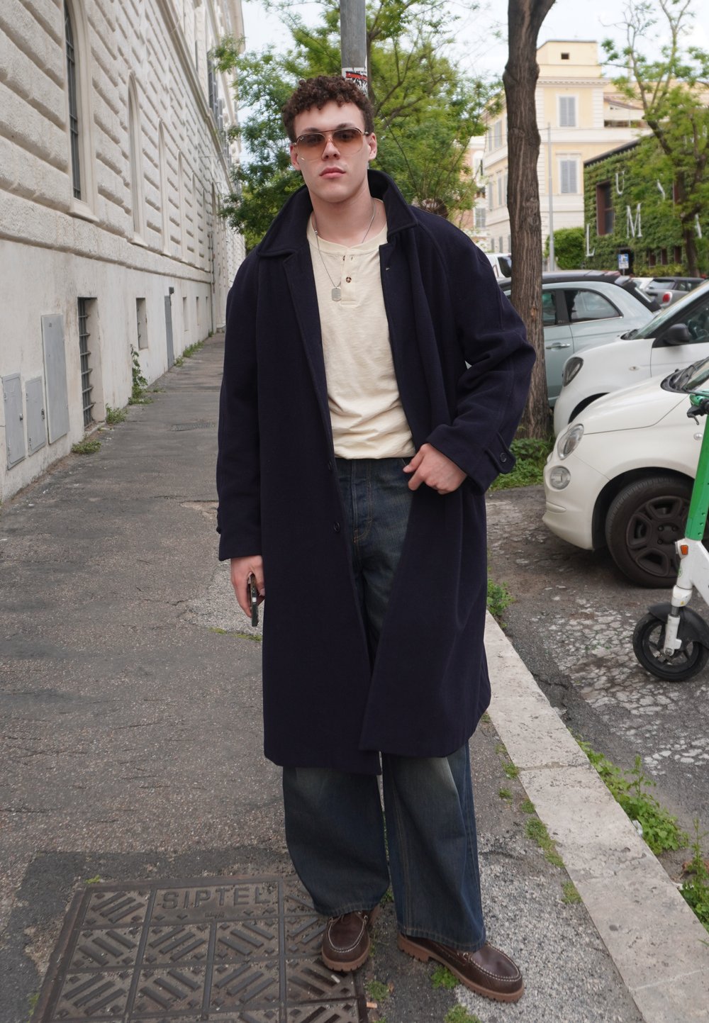 Um longo casaco azul-marinho sobre uma camisa creme, jeans de perna larga azuis e sapatos castanhos. Os acessórios incluem óculos de sol e um telemóvel na mão. Cenário urbano.