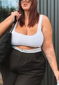 Top cropped branco, calças pretas de cintura alta com uma faixa baixa, e uma mala preta. Cabelo castanho longo e liso, tatuagens visíveis.