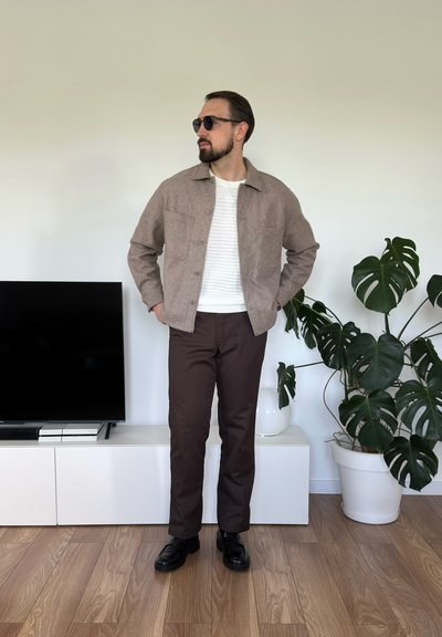 Chaqueta de mezcla de lana marrón con dos bolsillos delanteros, suéter blanco texturizado, pantalones marrones y zapatos negros contra un fondo neutro.