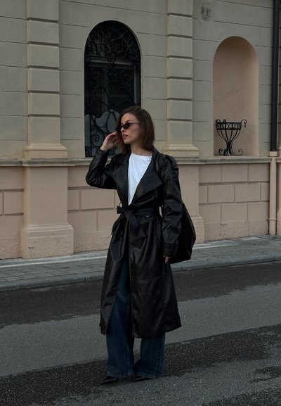 Mujer con abrigo largo de cuero negro, camisa blanca, jeans azules y gafas de sol parada en una calle frente a un edificio beige con ventanas arqueadas.