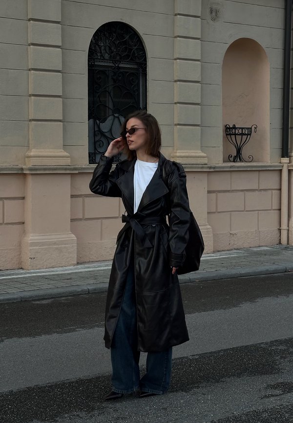 Donna con trench nero di pelle, camicia bianca, jeans blu e occhiali da sole in piedi su una strada di fronte a un edificio beige con finestre ad arco.