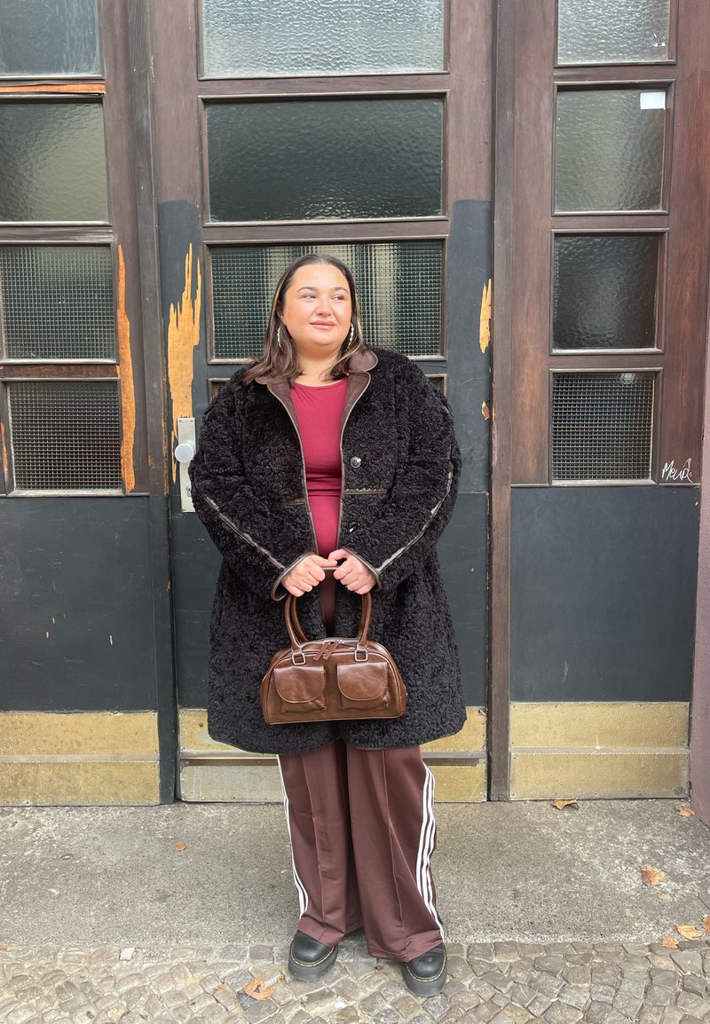 Black, fluffy sheepskin coat with brown leather accents, worn over a burgundy top. Brown handbag with two pockets, paired with brown trousers featuring white stripes.