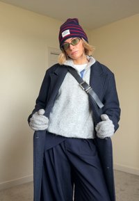 Manteau en laine bleu marine porté sur un pull gris, complété par un bonnet rayé bleu marine et rouge, des gants gris, et un pantalon oversize bleu marine.