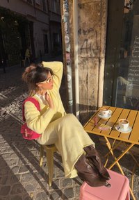 Conjunto amarelo tricotado, botas castanhas pontiagudas, mala rosa texturada, sentado numa mesa amarela com chávenas de café e um batom em cima.